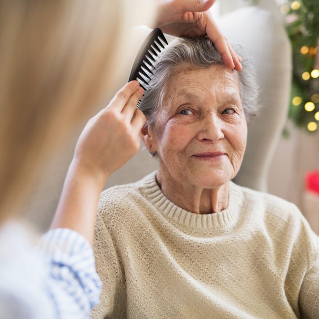 A health visitor combing hair of senior woman at home at Christmas time. An unrecognizable health visitor combing hair of senior woman sitting on a sofa at home at Christmas time.