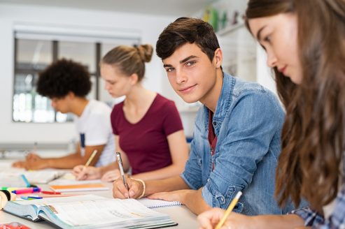 Guy studying with friends at college Portrait of happy college young man studying with classmates in library. Group of multiethnic friends studying together while sitting in university classroom. High school guy looking at camera while sitting in a row with girls at desk.
