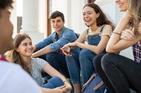 Teenager friends sitting together and laughing Group of happy young friends sitting in college campus and talking. Cheerful group of smiling girls and guys feeling relaxed after university exam. Excited millenials laughing and having fun outdoor.