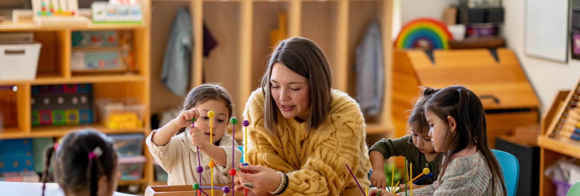 A teacher sits with a small group of young students at a desk, as they play with various building materials.  The children are each dressed casually and appear engaged in the task.
